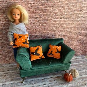 May include: A doll with blonde hair wearing a grey sweater stands beside a miniature dark green velvet sofa. The sofa features two orange throw pillows decorated with black witch hat and white star patterns. Two small fabric pumpkins sit nearby.