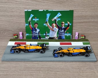 Könnte beinhalten: Ein gelbes und blaues Modell eines Rennwagens auf einer Miniaturrennstrecke mit einer karierten Ziellinie. Das Auto trägt den Schriftzug "Alteryx" an der Seite. Hinter der Rennstrecke ist ein Foto von drei Personen zu sehen, die Trophäen halten.