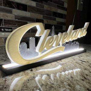 May include: A white illuminated sign with the word "Cleveland" in a stylized font. The sign features a cityscape silhouette behind the text.