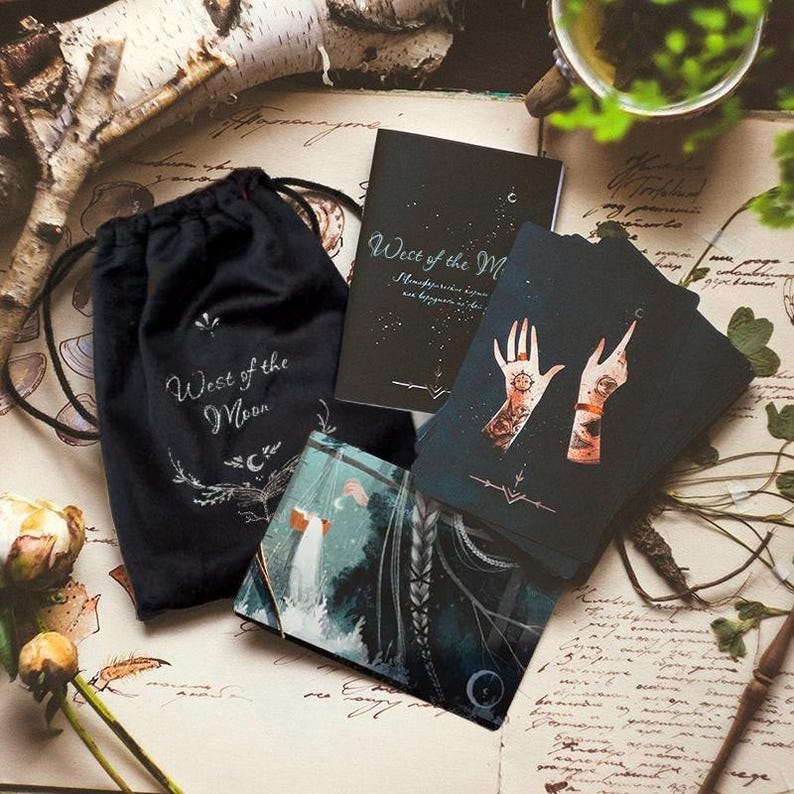 Puede incluir: Una bolsa de terciopelo negro con las palabras "West of the Moon" impresas en ella. La bolsa est&aacute; junto a una baraja de cartas de tarot con fondo negro e ilustraciones de manos. Las cartas tienen las palabras "West of the Moon" impresas en ellas.