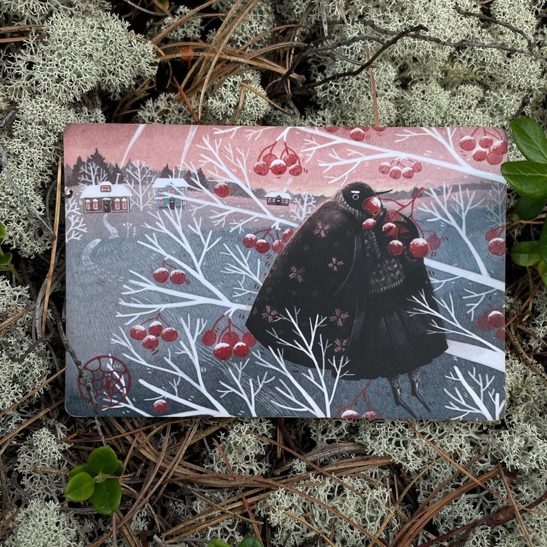 Puede incluir: Un p&aacute;jaro negro con una capa negra y una bufanda roja se encuentra en un bosque nevado. El p&aacute;jaro sostiene bayas rojas en su pico. El fondo es un bosque nevado con casas en la distancia.