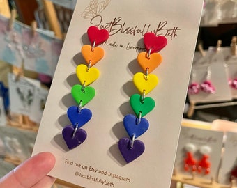 Könnte beinhalten: Regenbogen-Herz-Ohrringe aus Polymer Clay. Die Ohrringe sind in den Farben des Regenbogens, mit einem roten Herz oben und einem violetten Herz unten. Die Ohrringe befinden sich auf einer weißen Karte mit dem Text "Just Blissfully Beth Made in Liverpool" und "Find me on Etsy and Instagram @justblissfullybeth".