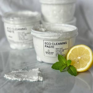 May include: White eco-cleaning paste in a clear plastic container with a label. The label reads "ECO CLEANING PASTE" and lists ingredients. A lemon half and mint sprig are next to the container. A small amount of paste is spread out.