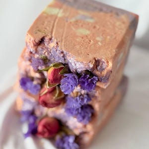 May include: Handmade soap bars stacked, with a textured, rustic look. The soap has a peach and lavender colour scheme, decorated with dried purple flowers and rosebuds. The soap is square in shape.