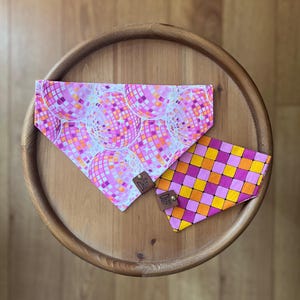 May include: Two dog bandanas, one with a pink and orange disco ball pattern and the other with a pink and orange checkered pattern.