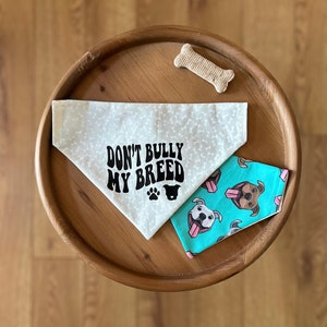 May include: Two dog bandanas and a bone-shaped dog treat on a wooden tray. One bandana is cream-colored with a bone pattern and the text "DON'T BULLY MY BREED". The other is turquoise with a dog face pattern.