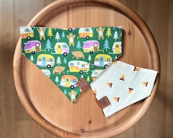 May include: Two dog bandanas on a wooden tray. One bandana is green with colorful camper vans, bears, and trees. The other is cream-colored with campfire graphics. Both have leather tags with the brand logo.