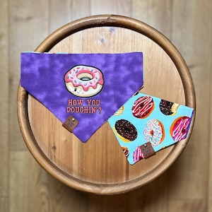 May include: Two dog bandanas. One is purple with a donut graphic and the text "HOW YOU DOUCHIN?". The other is light blue with a donut pattern. Both have a small leather tag.