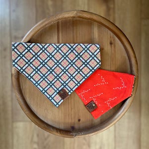May include: Two dog bandanas on a wooden surface. One bandana has a tan, black, and red plaid pattern. The other is bright red with a white dotted design. Both have a small leather tag.