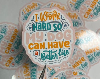 May include: A pink and white sticker with the text "I work hard so my dog can have a better life" in colorful, stylized lettering. The sticker is surrounded by other similar stickers.