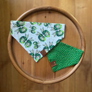 May include: Two dog bandanas. One is light blue with a repeating pattern of green bows with red and white accents. The other is green with a white polka dot pattern. Both have a brown leather tag.