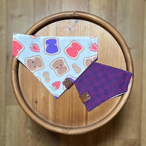 May include: Two dog bandanas. One features a light blue background with cartoon images of peanut butter and jelly sandwiches, strawberries, and peanuts. The other is a purple and red checkered pattern. Both have leather tags.