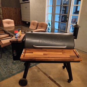 May include: Black Traeger wood pellet grill with a wooden cutting board on top. The grill has wheels and is on a concrete patio.