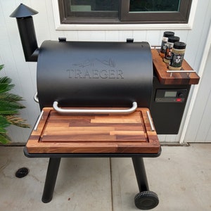 May include: Black Traeger wood pellet grill with a wood cutting board and a side shelf with three spice jars. The grill has a digital control panel and a black metal handle.