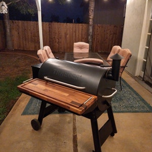 May include: Black Traeger wood pellet grill with a wooden cutting board on the side. The grill is on a patio with string lights overhead.