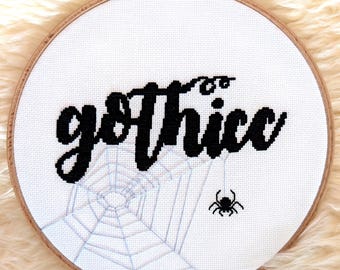 May include: A cross-stitch embroidery hoop with the word "gothicc" in black script. A spider and web are stitched below the text. The hoop is set against a fluffy, cream-colored background.