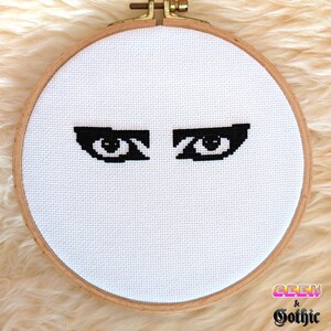 May include: A cross-stitch embroidery hoop featuring a pair of black eyes on a white background. The embroidery is set in a wooden hoop with a brass clasp. The text "Geek & Gothic" is visible in the lower right corner.