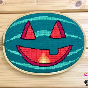 May include: Cross-stitch embroidery of a jack-o'-lantern face in a wooden hoop. The design features a teal background with a red and orange smiling face. The text "GEEK & Gothic" is in the bottom right corner.