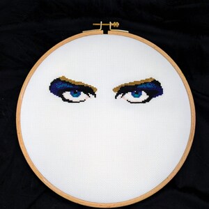 May include: A cross-stitch embroidery piece featuring a pair of detailed eyes with bright blue irises, dark blue and gold eyeshadow, and black eyeliner, centered on a white fabric canvas held within a circular wooden embroidery hoop.