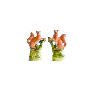 Può includere: Due statuette in ceramica raffiguranti scoiattoli arancioni appollaiati su basi verdi testurizzate. Ogni figura di scoiattolo presenta una finitura lucida, una coda curva e un piccolo ritaglio circolare al centro del corpo.