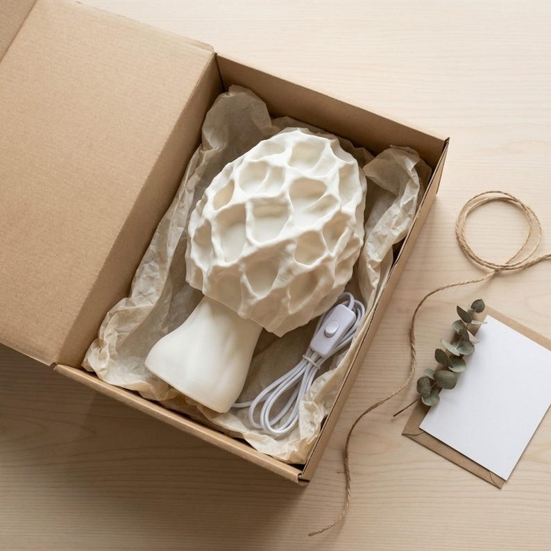 May include: A white mushroom-shaped lamp with a textured cap and a smooth stem, inside a cardboard box. The lamp has a white cord and switch. The box is lined with brown paper. A blank card and eucalyptus sprig are on the wooden surface.