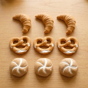 Könnte beinhalten: Eine Ansammlung von neun Miniatur-Backwaren, angeordnet in drei Reihen. Die oberste Reihe zeigt drei goldbraune Croissants, die mittlere Reihe enthält drei Brezeln mit weißen Salzdetails und die untere Reihe zeigt drei runde Brötchen.
