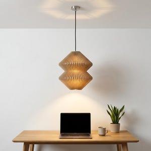 Könnte beinhalten: Eine Pendelleuchte mit einem hellbraunen, gerippten, zweistufigen geometrischen Lampenschirm, der an einem dünnen schwarzen Kabel hängt. Die Lampe strahlt von innen ein warmes, sanftes Licht aus, das nach unten auf die Fläche leuchtet.