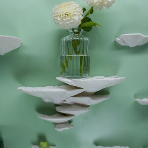 May include: A clear glass vase with two white dahlias sits on a stack of white cloud-shaped shelves against a light green wall. A small green frog figurine rests on a cloud shelf. Decorative wall art.