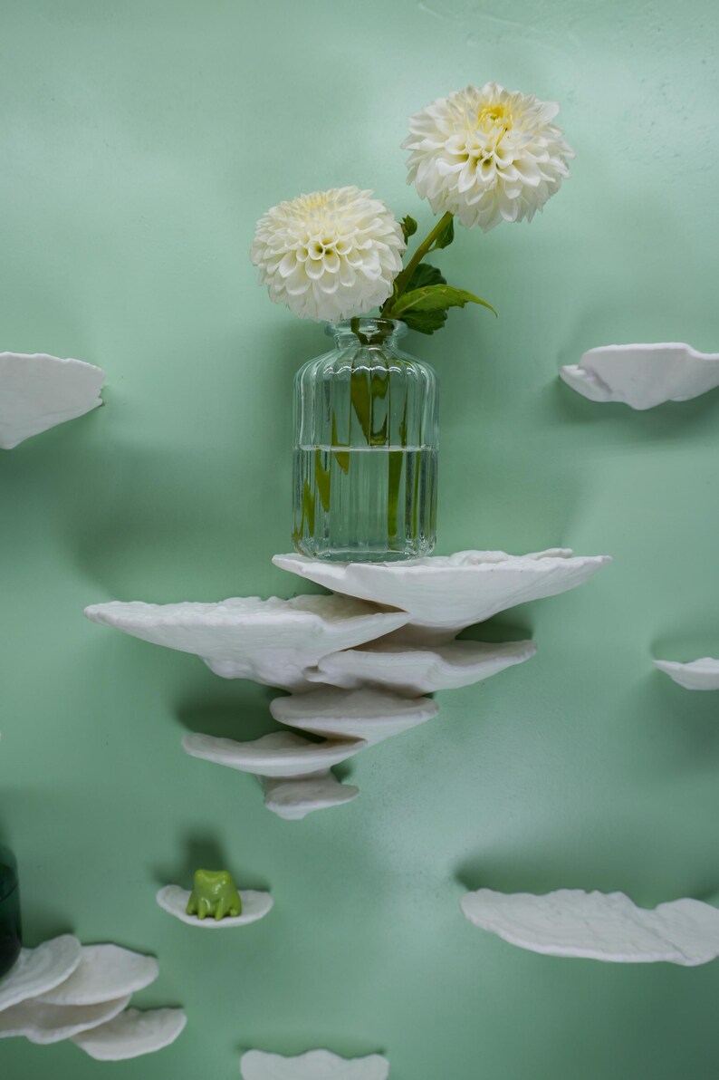 May include: A clear glass vase with two white dahlias sits on a stack of white cloud-shaped shelves against a light green wall. A small green frog figurine rests on a cloud shelf. Decorative wall art.