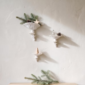 Könnte beinhalten: Drei weiße Wandregale aus Keramik in Pilzform. Die Regale sind vertikal angeordnet und mit kleinen Dekorationsgegenständen wie Tannenzweigen, einem Holzstern, einer klaren Glaskugel und einem kleinen Weihnachtsbaum aus Holz bestückt.
