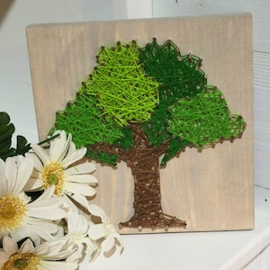 May include: A handmade string art piece depicting a tree. The tree features a brown trunk and a vibrant green and light green canopy, all created with string on a light wood background. Small pink accents are visible.