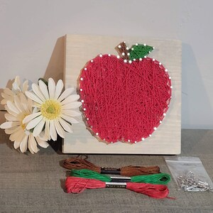 May include: A red string art apple on a wooden board with white daisy flowers and embroidery floss. The apple is made with red thread and white nail heads. The apple has a green and brown stem.
