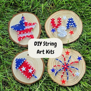 May include: Four DIY string art kits on wooden slices, featuring patriotic designs. The designs include a star, the letters "USA", a firework, and a burst of red, white, and blue string art. The kits are displayed on a bed of green grass.