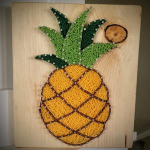 May include: A handmade string art pineapple on a wooden board. The pineapple features yellow and brown string, with green leaves. The design is accented with small silver nail heads. A small wooden circle is on the upper right corner.