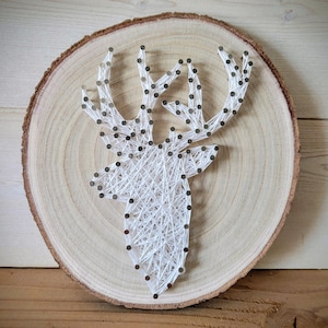 May include: A white string art deer head design on a round wooden slice. The deer head is created with white string wrapped around nails hammered into the wood.