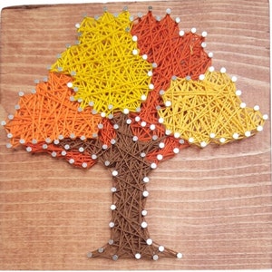 May include: A handmade string art tree on a wooden board. The tree features a brown trunk and colorful leaves in shades of yellow, orange, and red. The string is secured with small silver nails, creating a unique and textured artwork.