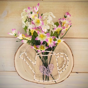May include: A wooden plaque with a wire-wrapped watering can design and a bouquet of pink and white flowers.