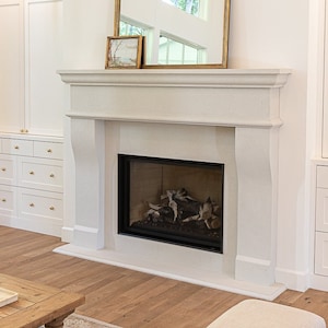May include: A white fireplace with a black firebox and a gold framed mirror above it. The fireplace is in a living room with white built-in cabinets and a white armchair.