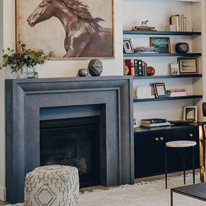 May include: A gray stone fireplace with a black mantel and a white rug in front of it. A brown and white patterned ottoman sits in front of the fireplace. A bookshelf with various books and decorative items is on the right side of the image.