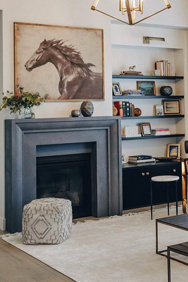 May include: A gray stone fireplace with a black mantel and a white rug in front of it. A brown and white patterned ottoman sits in front of the fireplace. A bookshelf with various books and decorative items is on the right side of the image.
