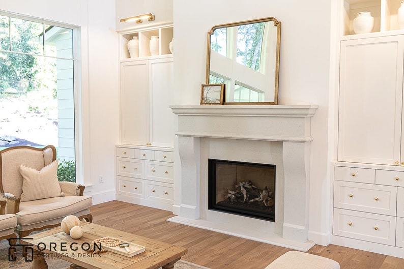 May include: A white fireplace with a black firebox and a gold framed mirror above it. The fireplace is in a living room with white built-in cabinets and a white armchair.