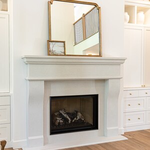 May include: A white fireplace with a black firebox and a gold-framed mirror above it. The fireplace is in a room with white walls and a wooden floor.
