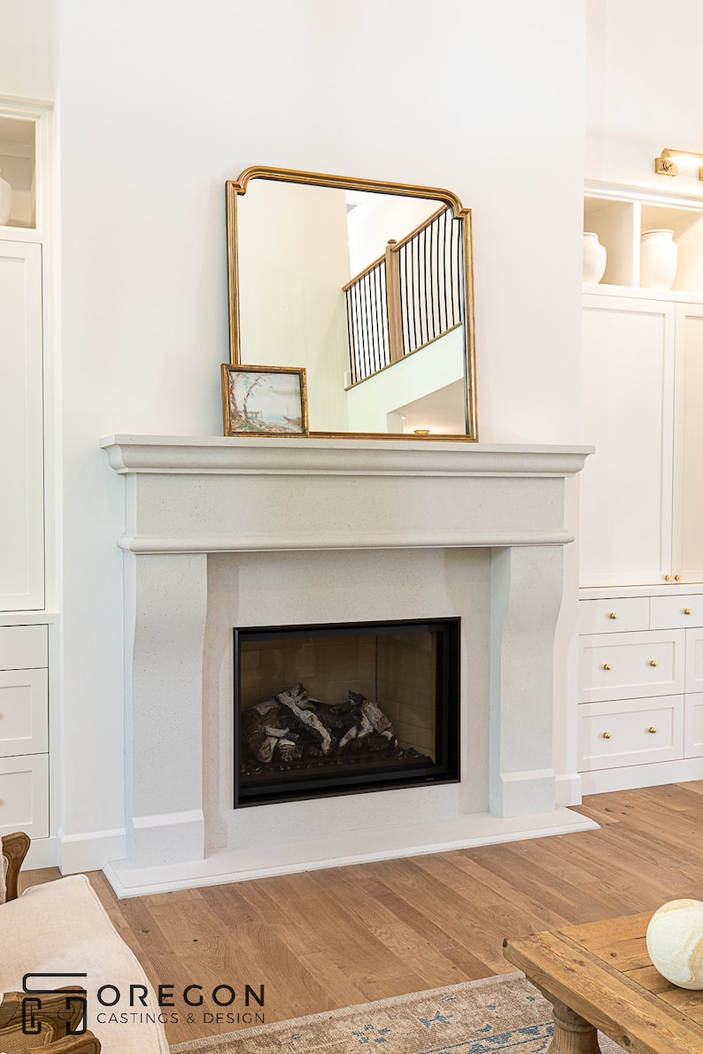 May include: A white fireplace with a black firebox and a gold-framed mirror above it. The fireplace is in a room with white walls and a wooden floor.