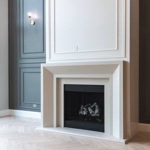 May include: A white fireplace with a black firebox and a wood-burning fire. The fireplace is set against a white wall with a dark gray wall to the left. The floor is a light wood herringbone pattern.