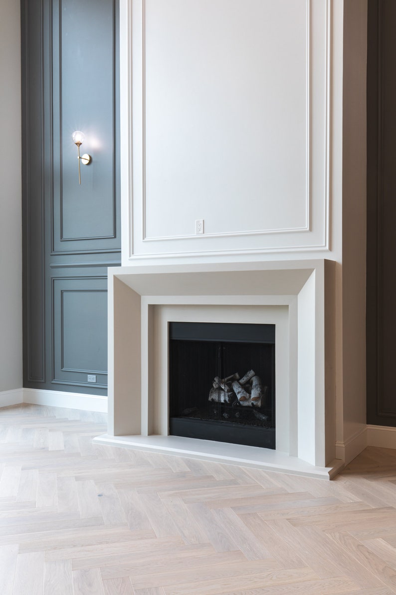 May include: A white fireplace with a black firebox and a wood-burning fire. The fireplace is set against a white wall with a dark gray wall to the left. The floor is a light wood herringbone pattern.