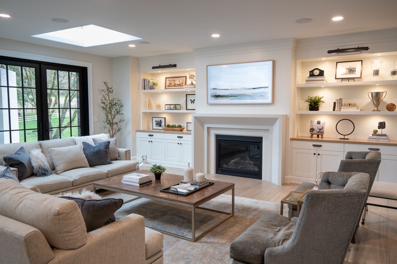 May include: A living room with a large sectional sofa, two armchairs, a coffee table, and a fireplace. The room has a skylight, large windows, and built-in shelves with decorative items.