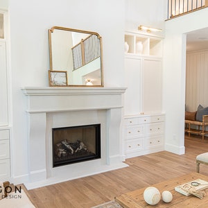 May include: A white fireplace with a gas log fire in a living room with white built-in cabinets and a large mirror above the fireplace.