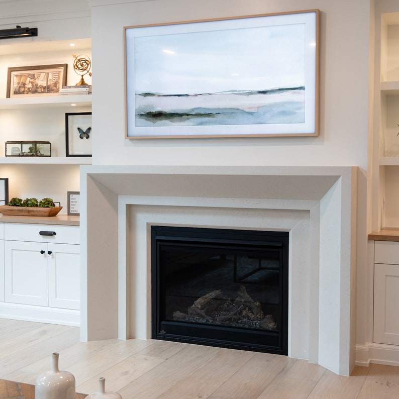 May include: A white fireplace with a black firebox and a framed abstract watercolor painting above it. The fireplace is in a modern living room with white walls and wood floors.