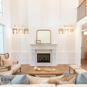 May include: A white living room with a fireplace, a large window, and a white built-in cabinet on each side of the fireplace. There are two beige armchairs in front of the fireplace and a wooden coffee table with a book and two white spheres on top. The room has a light wood floor and a white ceiling.