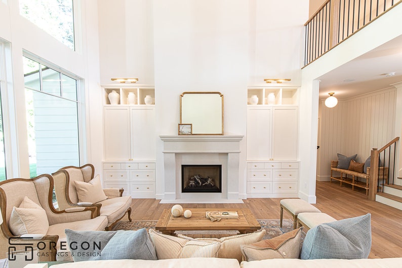 May include: A white living room with a fireplace, a large window, and a white built-in cabinet on each side of the fireplace. There are two beige armchairs in front of the fireplace and a wooden coffee table with a book and two white spheres on top. The room has a light wood floor and a white ceiling.